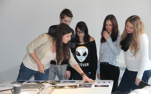 fünf Jugendliche stehen um verschiedene Plakatentwürfe, die auf einem Tisch liegen
